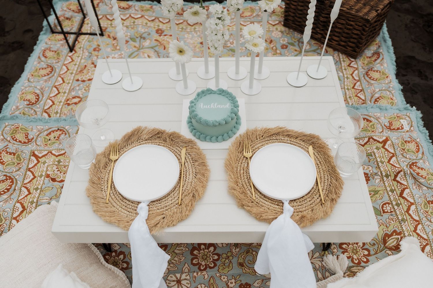 Place settings with white plates and woven placemats arranged on a table with orange floral patterned cloth.