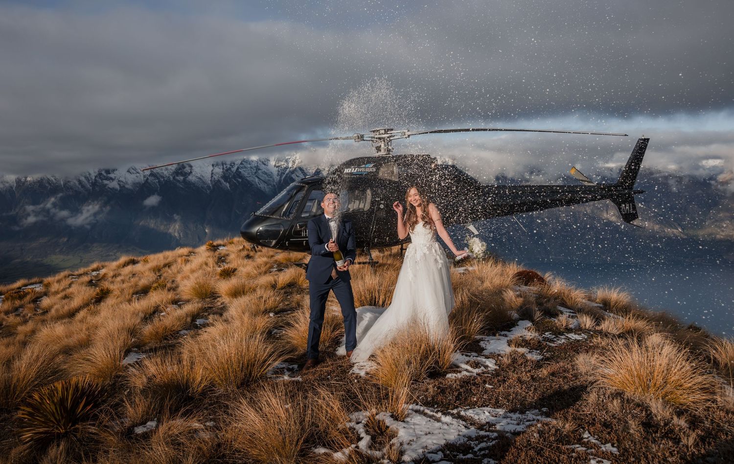 Scenic mountain-top wedding photo with helicopter and snow-capped peaks in the background.