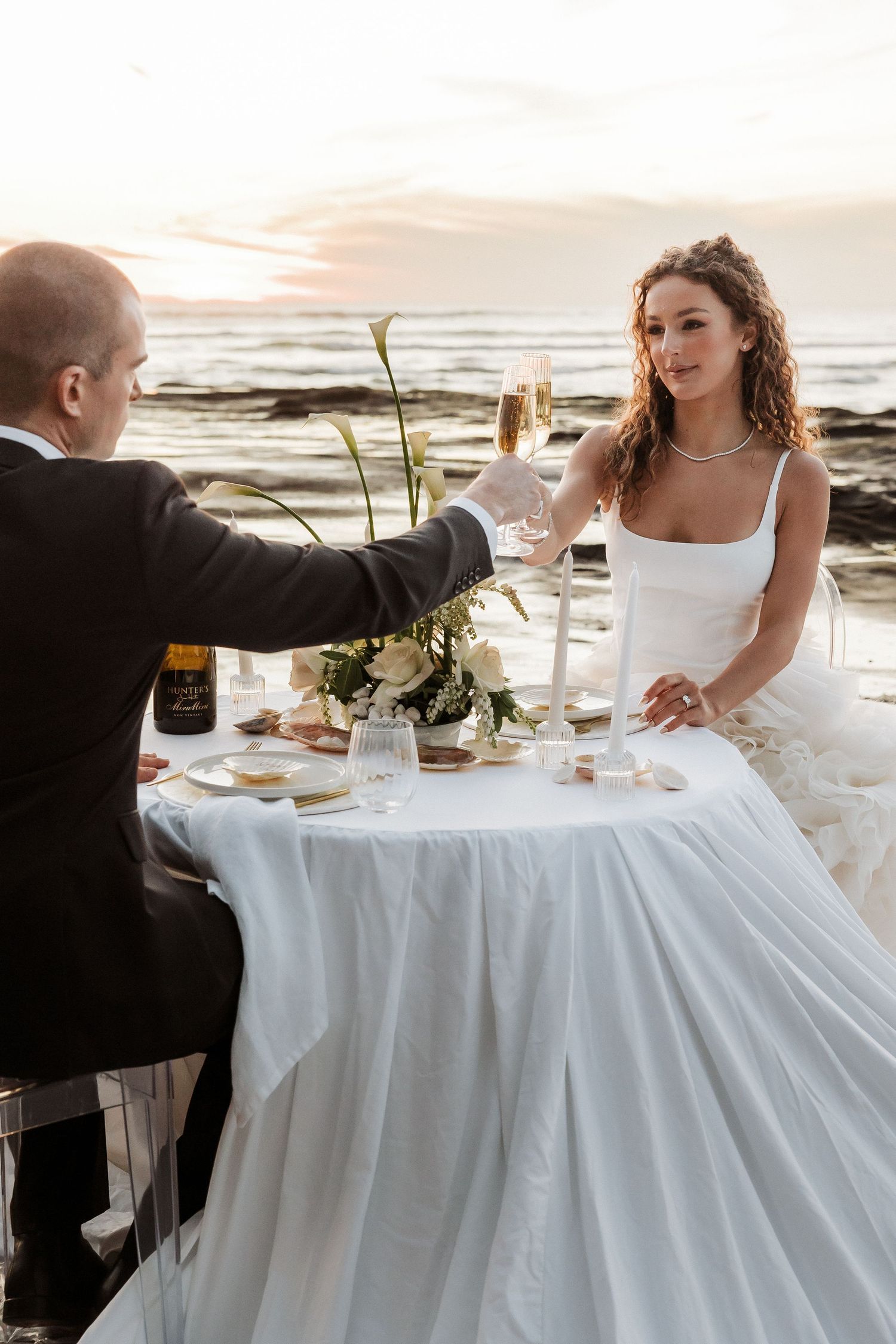 A romantic beachside wedding dinner setup with a white tablecloth and place settings at sunset.