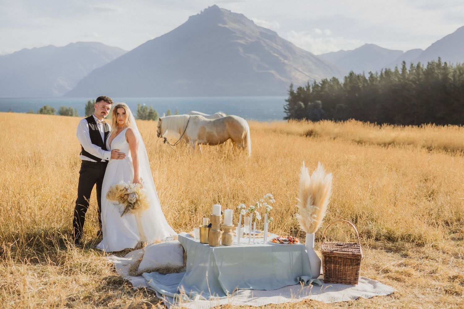 Two people share a romantic outdoor picnic with white horses and mountains in a golden field at sunset.
