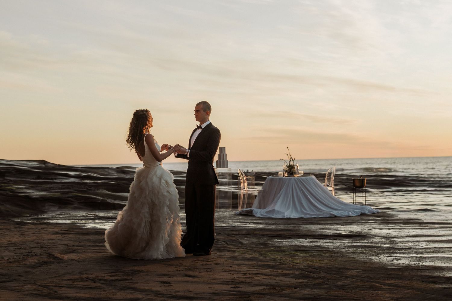 A couple in wedding attire sits on coastal rocks at sunset while waves crash against the shore in the background.