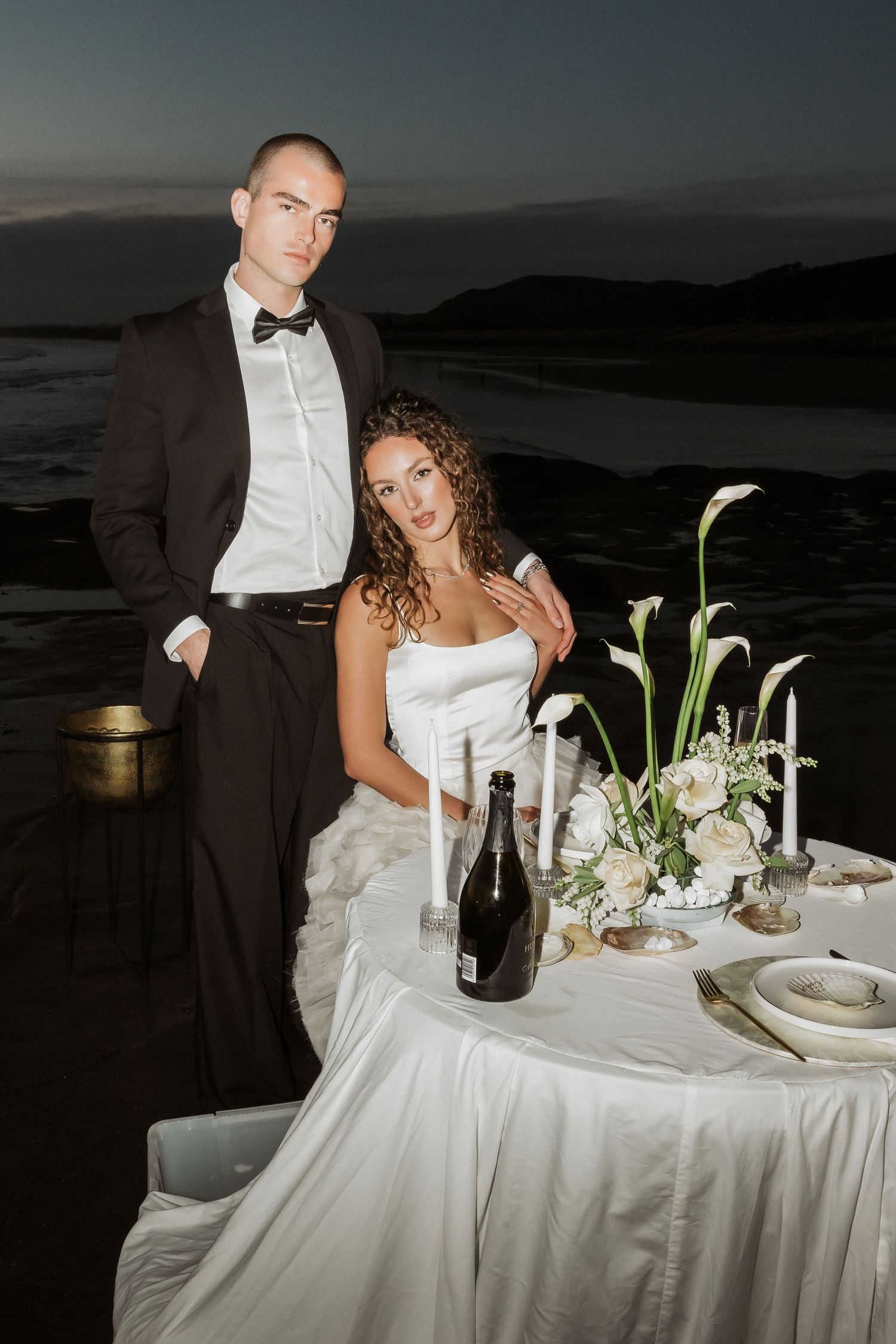 Romantic wedding reception table setting at sunset with white linens, flowers and candlelight overlooking ocean views.