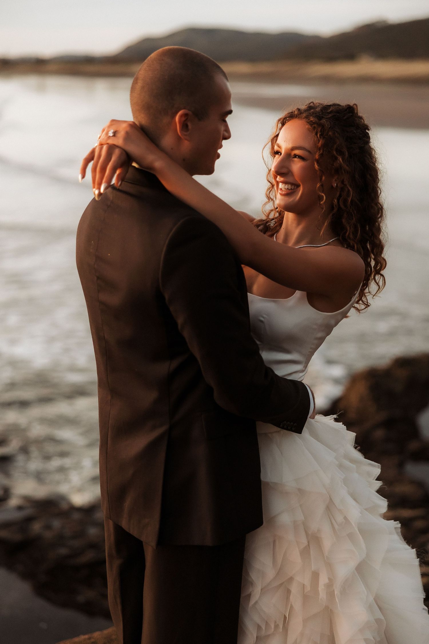 A couple shares an intimate moment on a rocky beach at sunset with dramatic lighting.