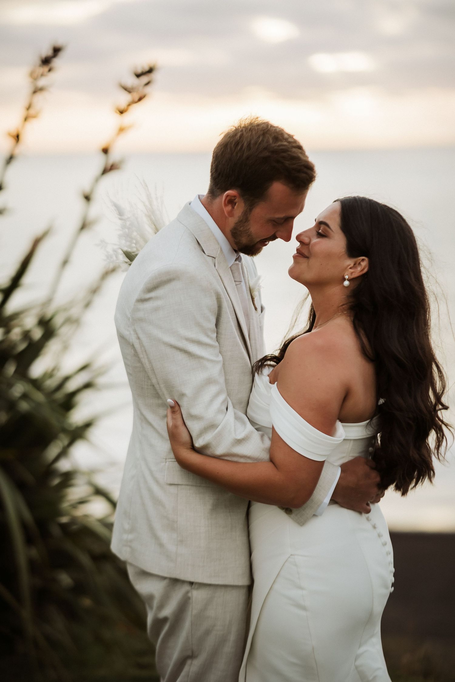 A romantic wedding couple embraces at sunset with tall grass silhouettes in the background.