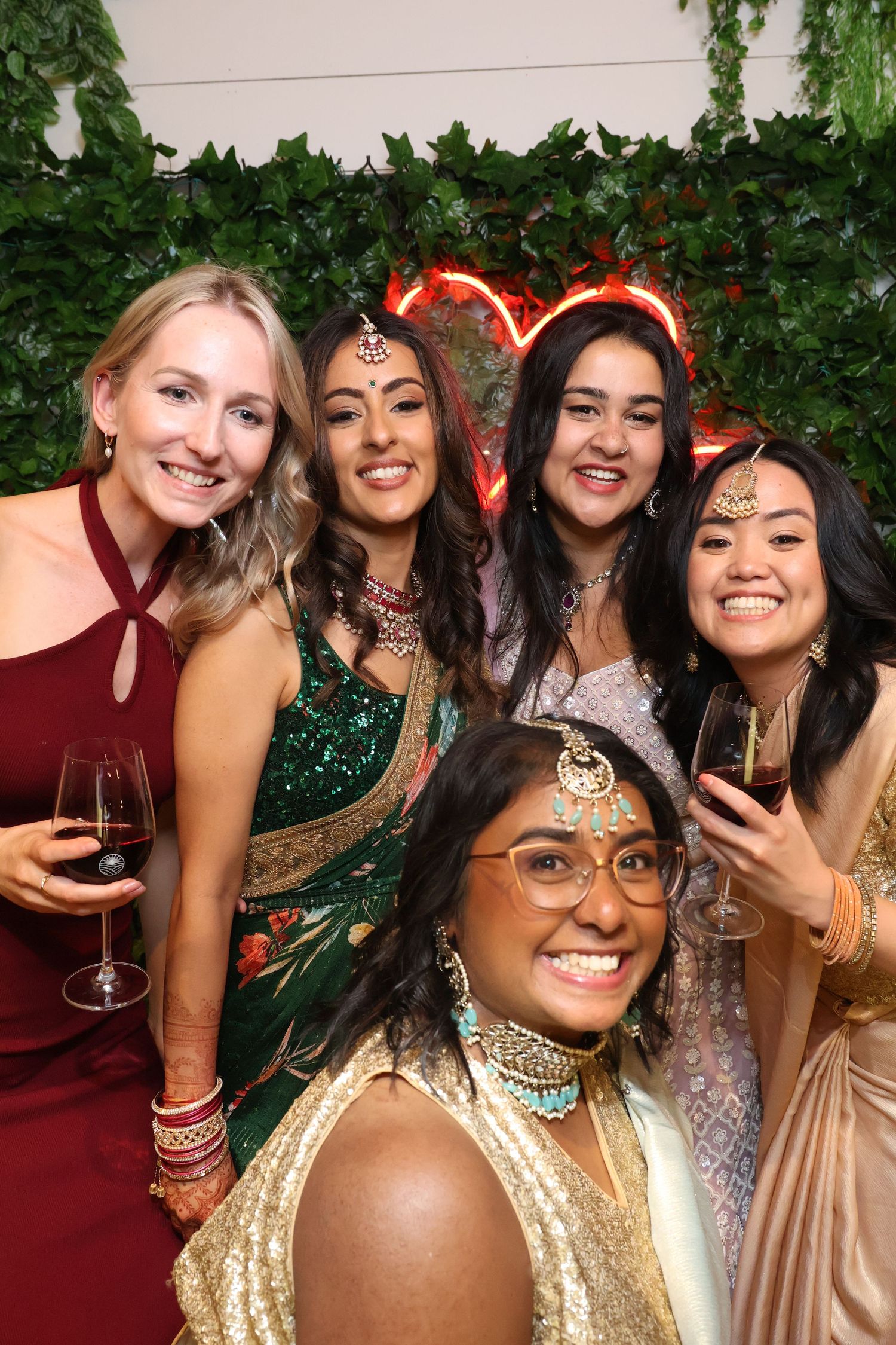 A group celebrates at a festive event wearing traditional Indian and Western attire against a leafy backdrop.