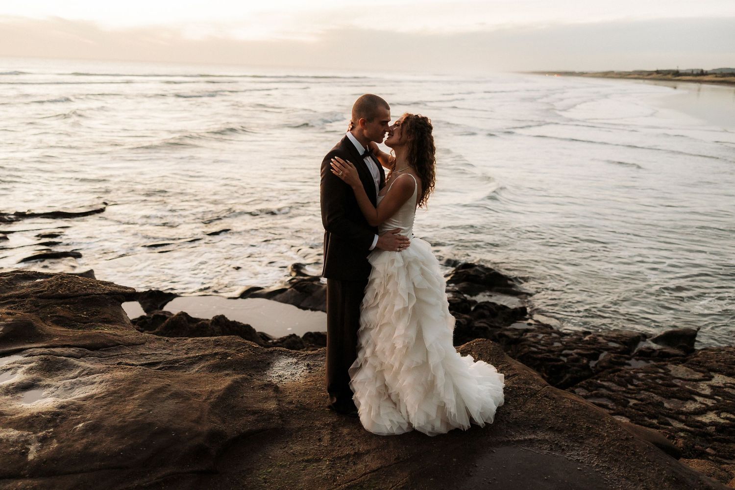 A couple embraces on rocky coastal cliffs during sunset as waves crash in the background.
