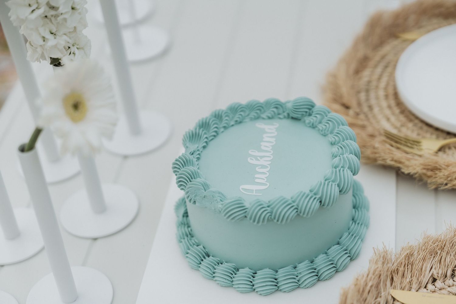 A mint green frosted cake with decorative piping sits on a white table with floral decor and natural elements.