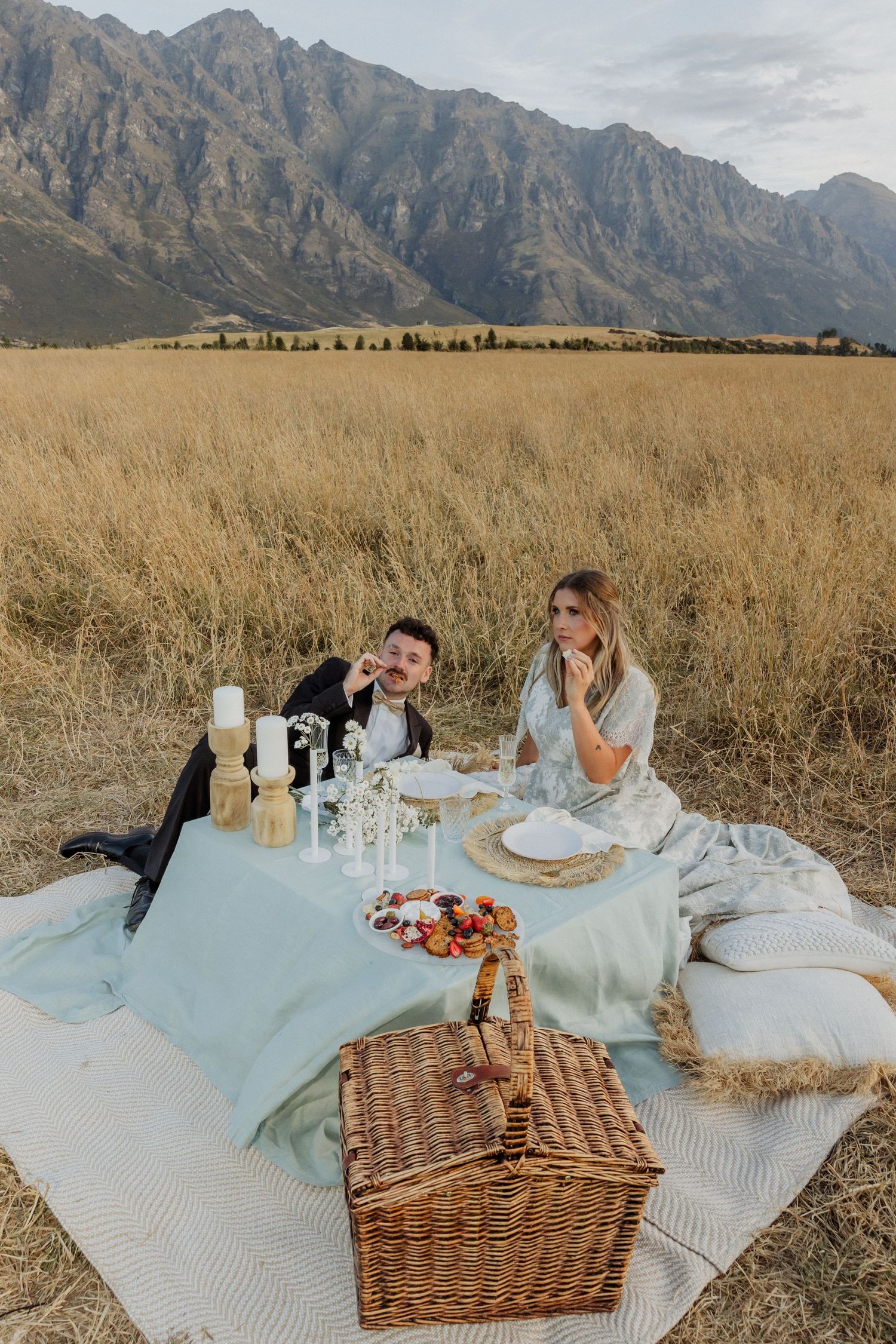 A romantic picnic setup with wicker basket and blanket against dramatic mountain backdrop in golden field.
