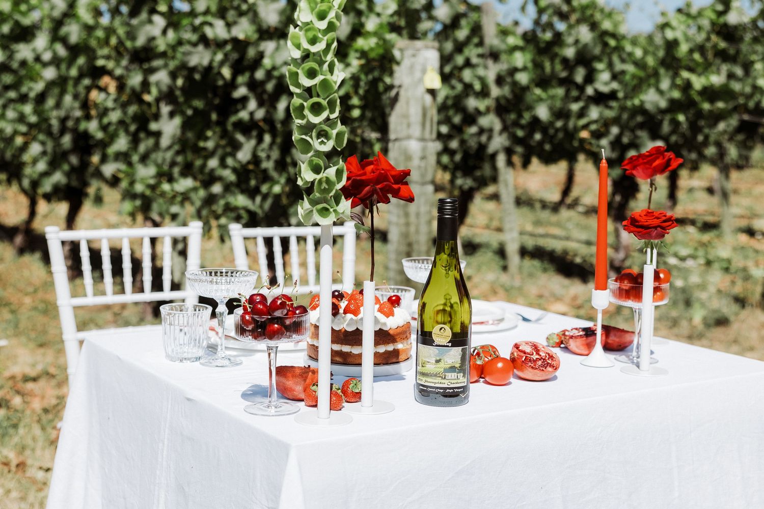 Elegant vineyard table setting with white tablecloth, wine glasses, red flowers and wine bottle in outdoor setting.