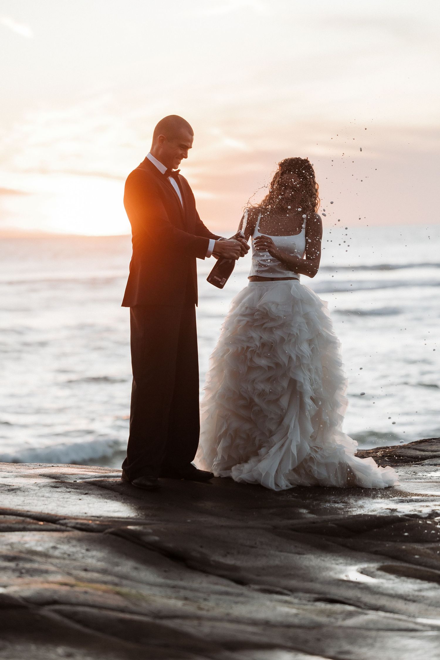 A couple celebrates their wedding at sunset on rocky coastal shores with waves crashing behind them.