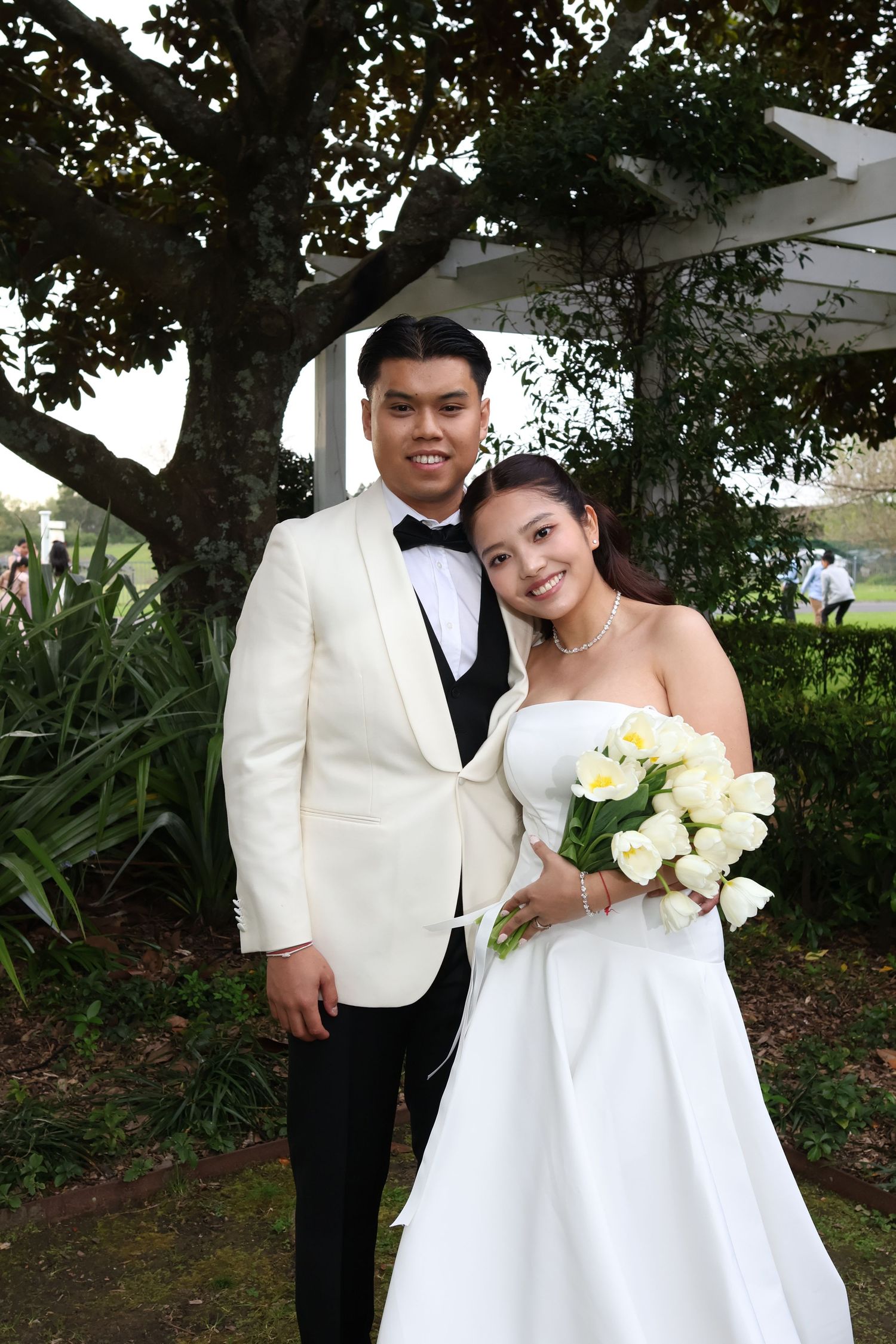A couple in formal wedding attire poses together outdoors under a tree canopy with white floral bouquet.