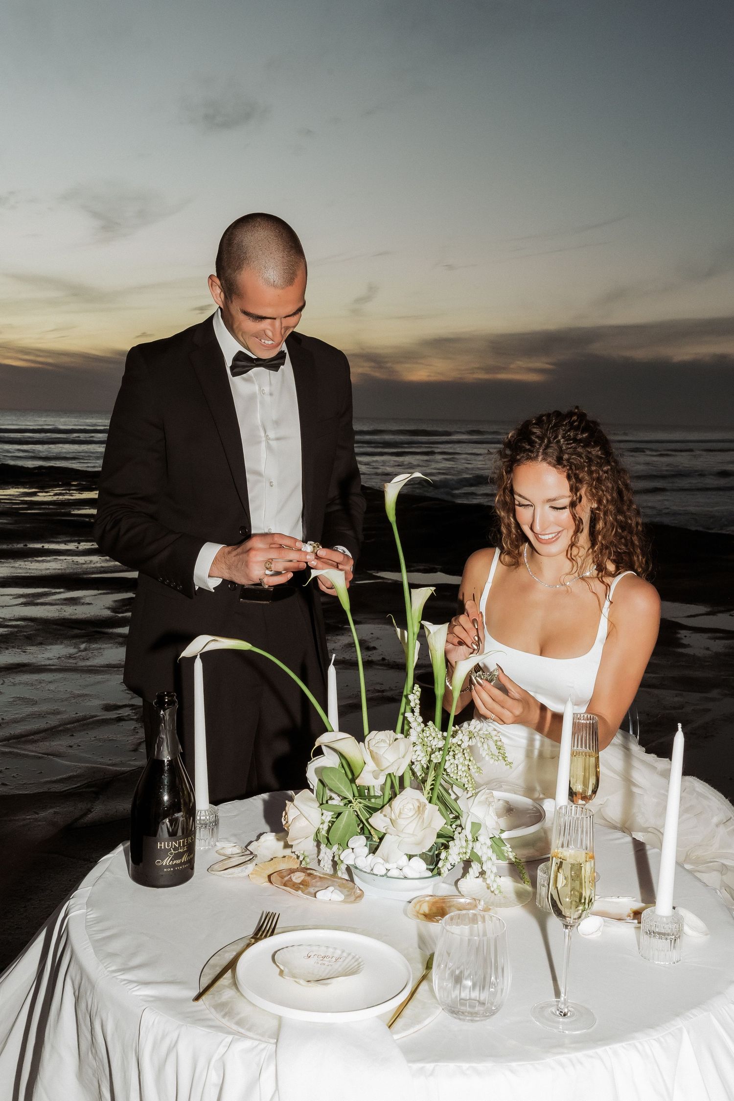 A romantic beach dinner setup with white linens, candles, and floral arrangements at sunset.