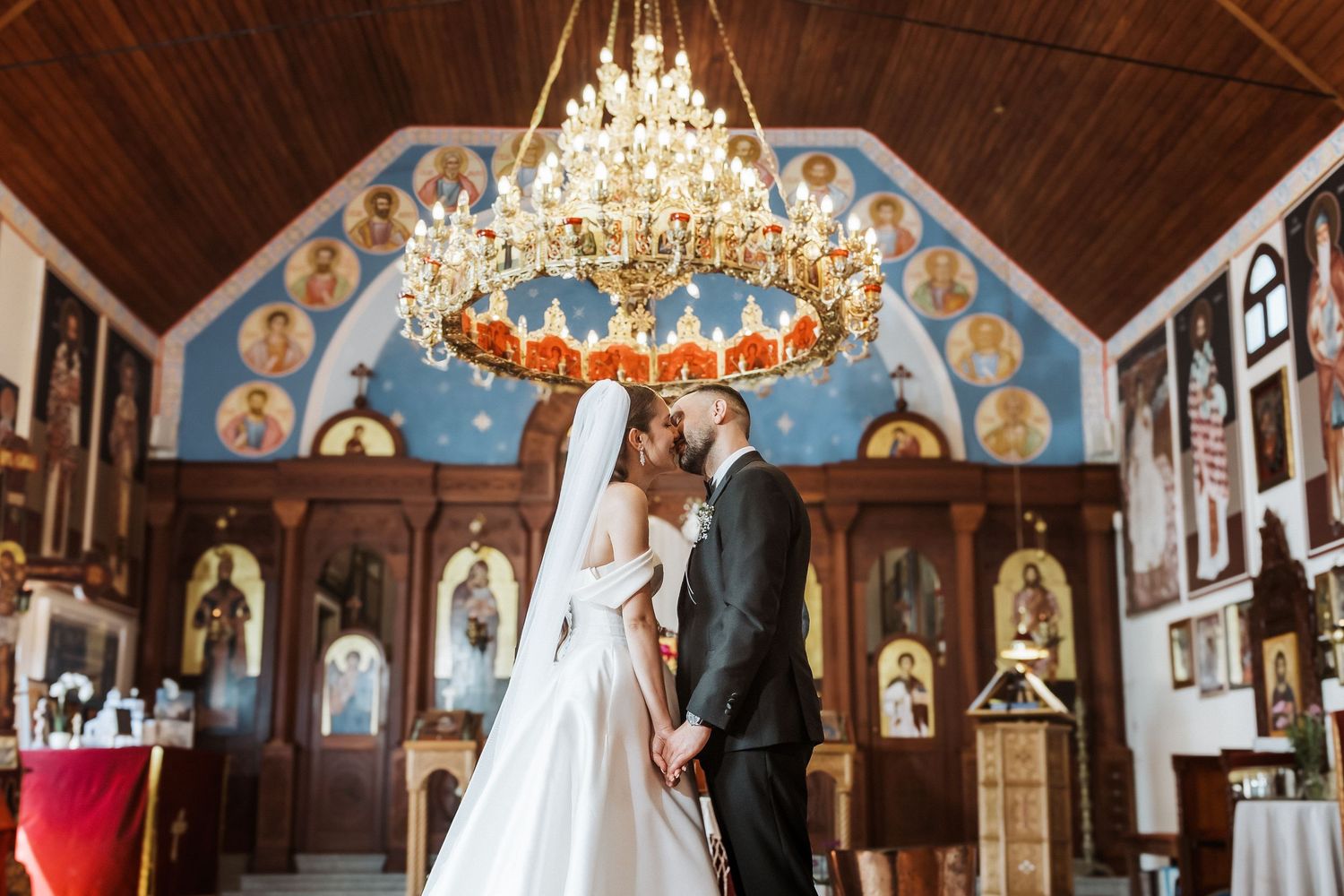 Wedding ceremony taking place in an ornate Orthodox church with wooden beams and chandeliers under a vaulted ceiling.