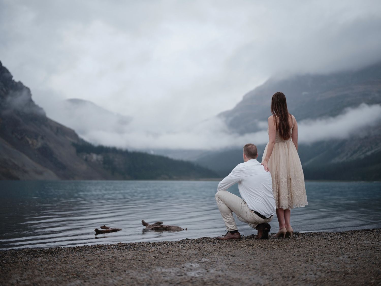 Couple standing at edge of misty mountain lake with dramatic foggy landscape and overcast sky in background. Canadian Rocky Mountains Banff