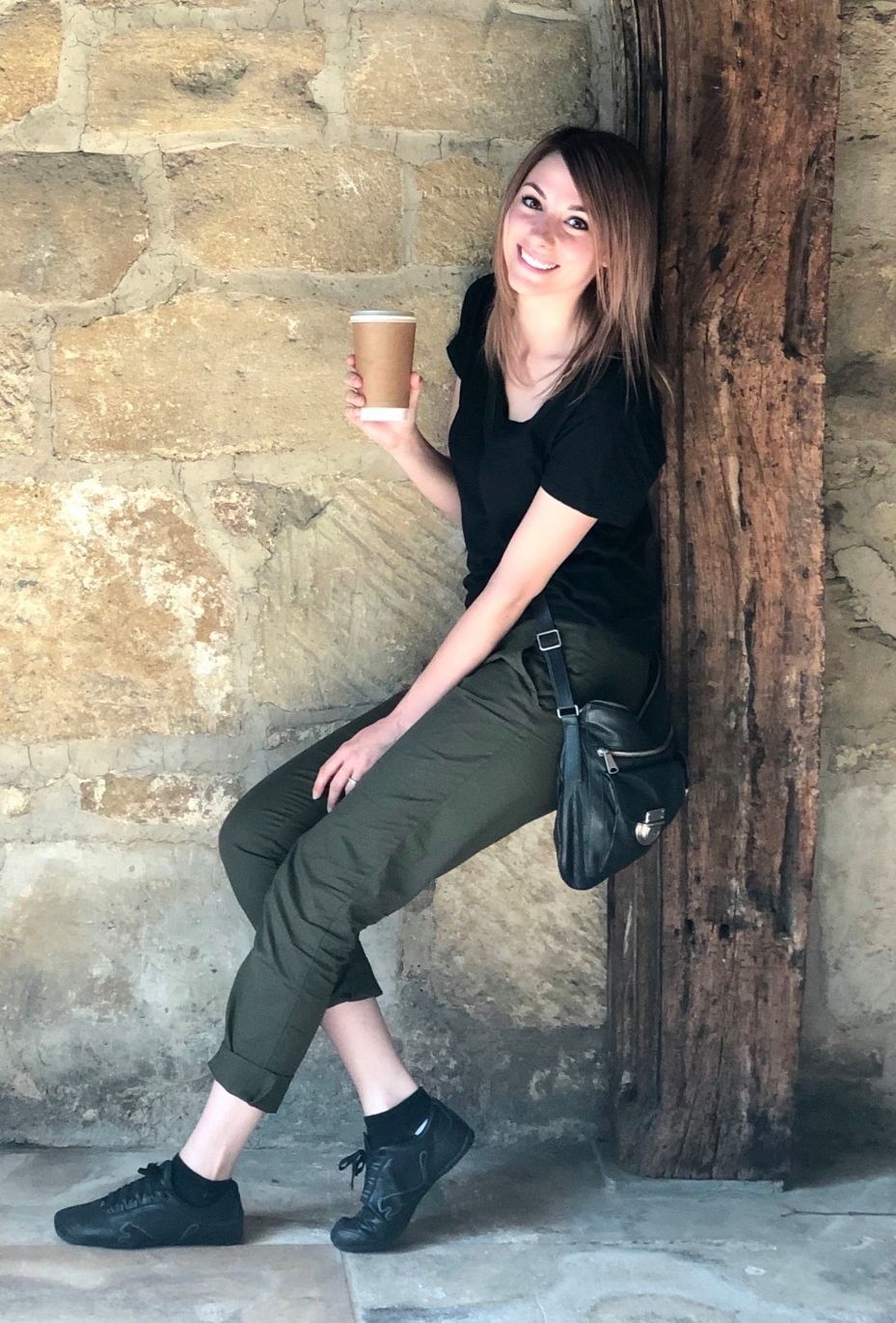 Kristen Lusk, wedding and elopement photographer at Sharko Studios holding a coffee cup, wearing black t-shirt and olive pants. Being cool