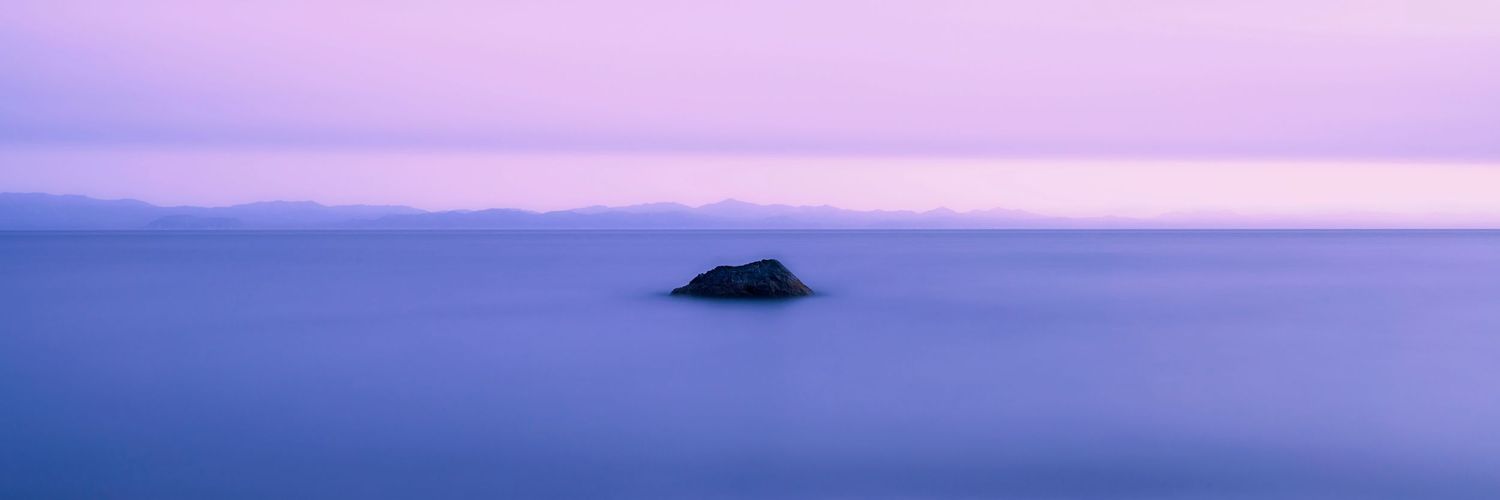 A solitary rock formation emerges from calm purple-tinted waters during a misty sunset over the ocean horizon.