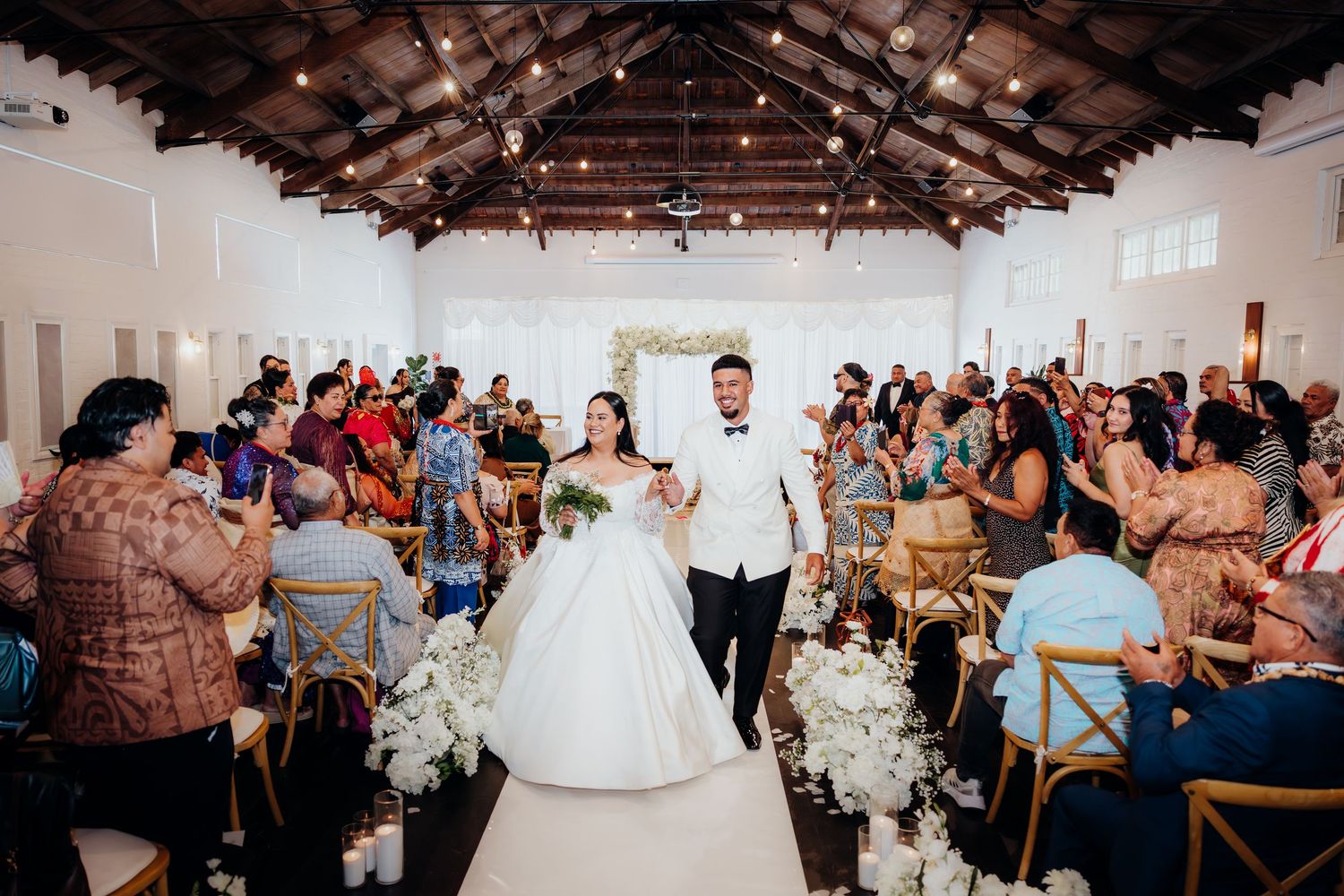 Wedding guests celebrate during a reception in a rustic venue with exposed wooden beams and white decor.