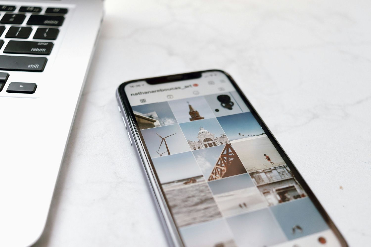 iPhone displaying grid of travel photos next to laptop keyboard on white surface.