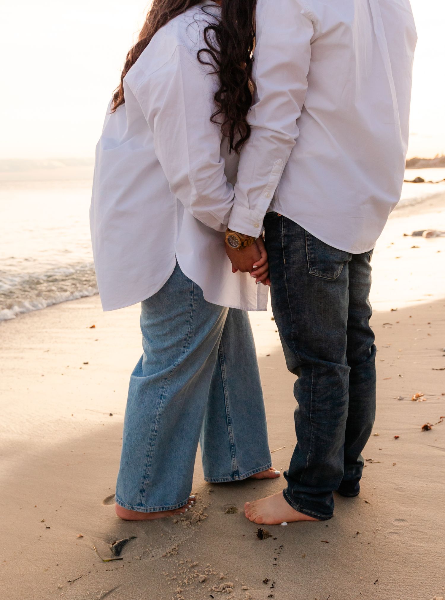 A couple wearing white shirts and jeans stands barefoot holding hands on a sandy beach at sunset.