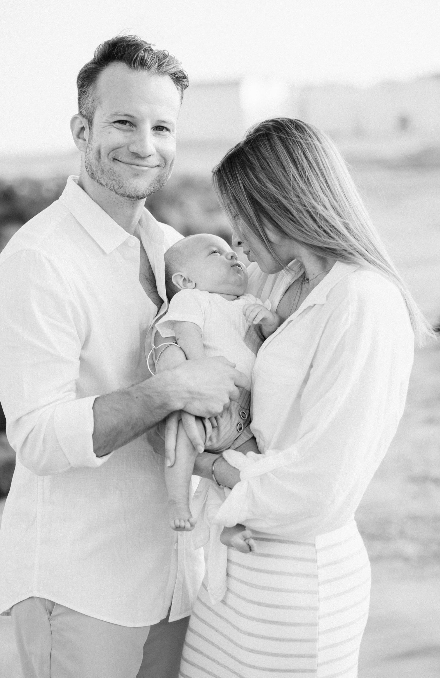 Parents holding newborn on the beach with ocean waves and sunset glow at Deal Beach in Monmouth County, NJ.