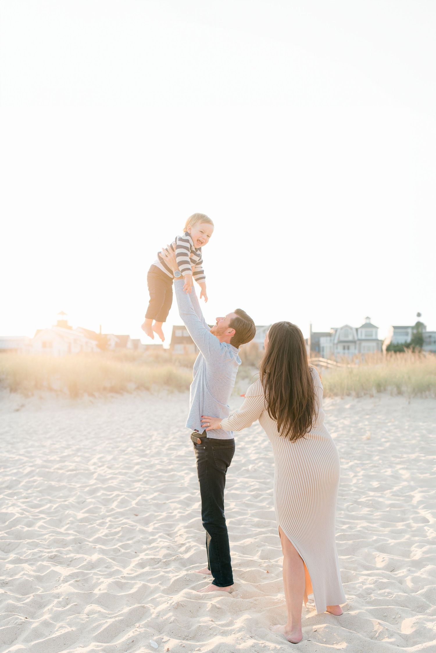 A man in a blue collared shirt lifts his son into the air while his wife stands behind them.