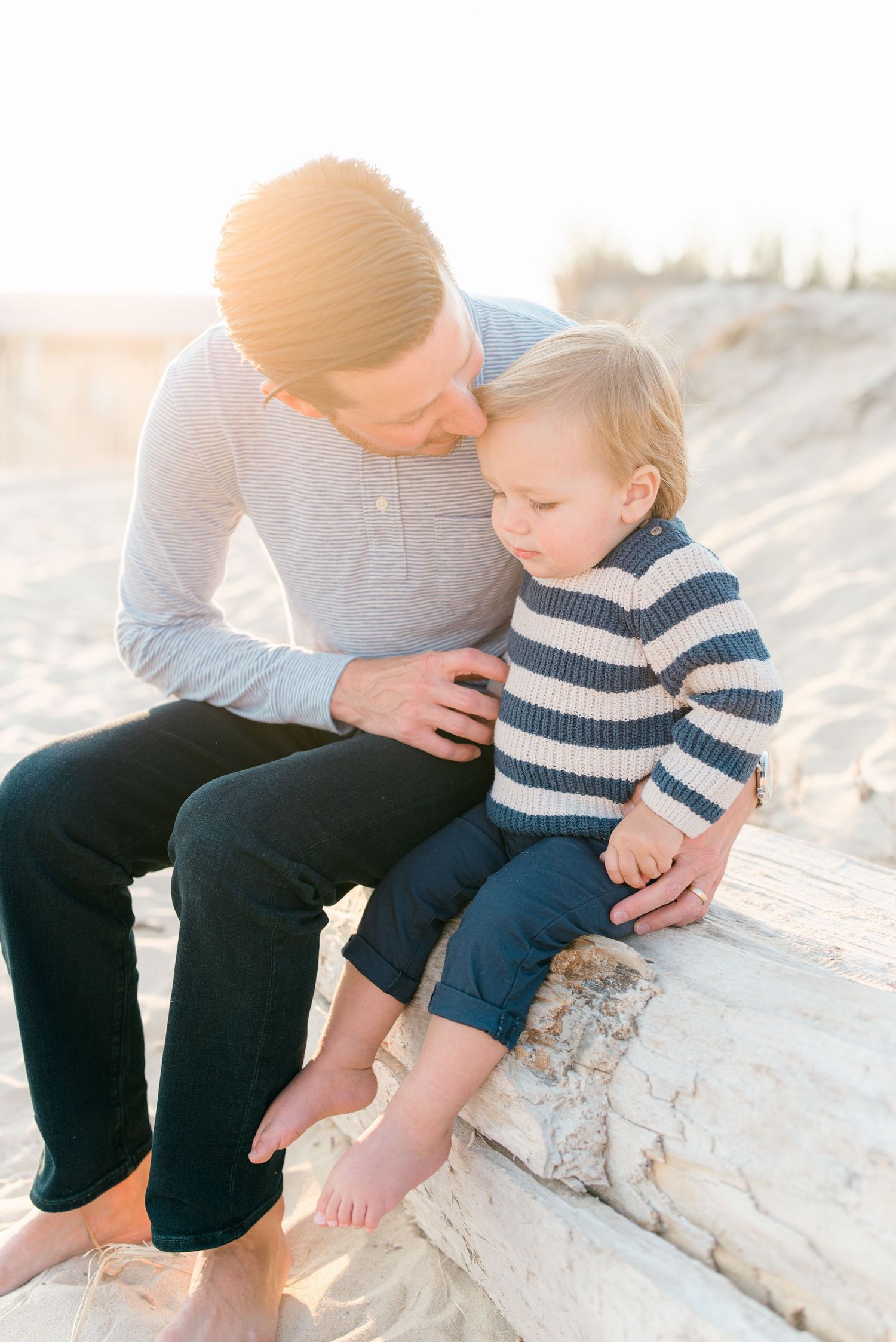 A father in a blue shirt and dark blue jeans kisses his son on the forehead while sitting on a piece of driftwood on the beach.