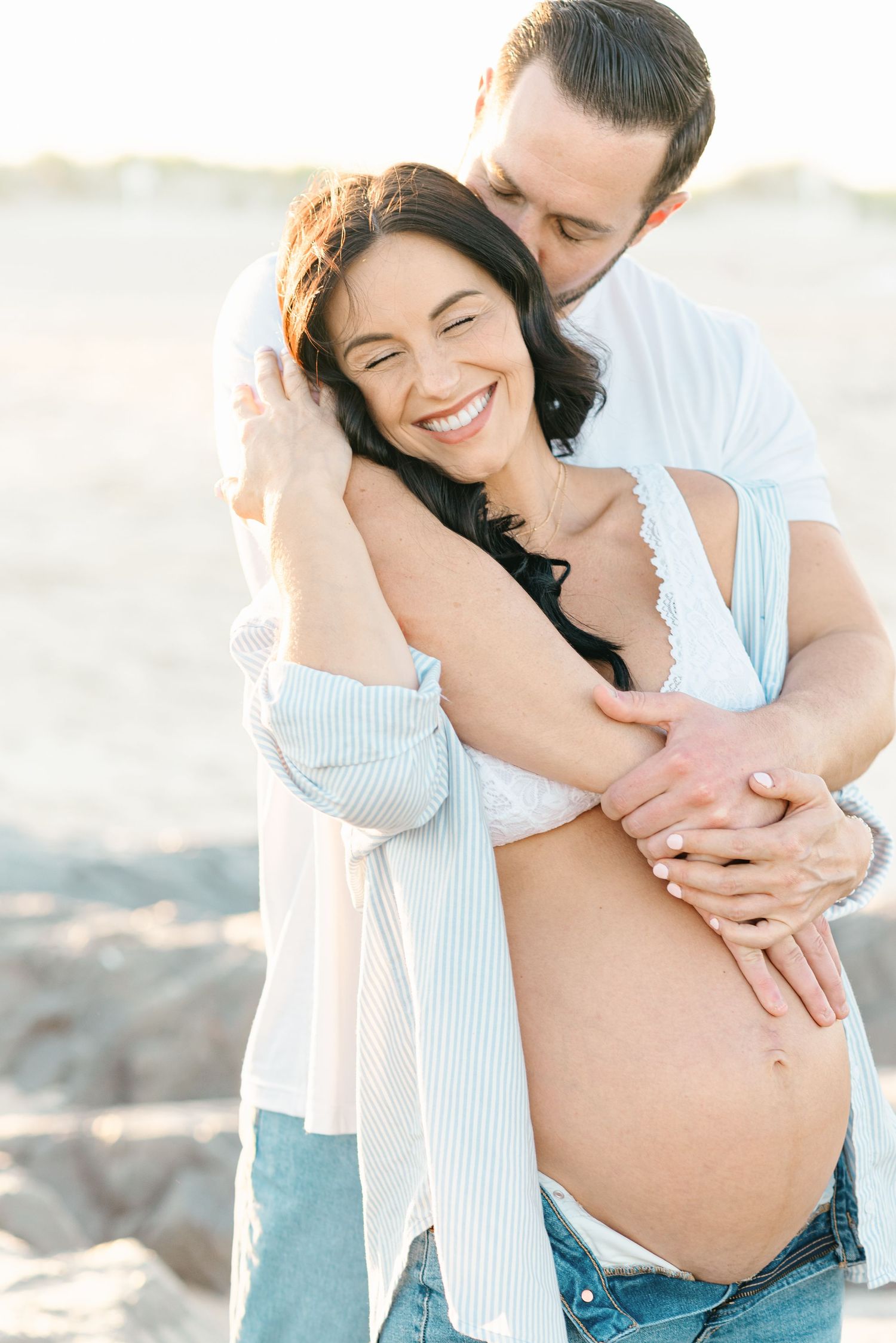 A man in a white shirt has his arms around a woman with a pregnant belly.
