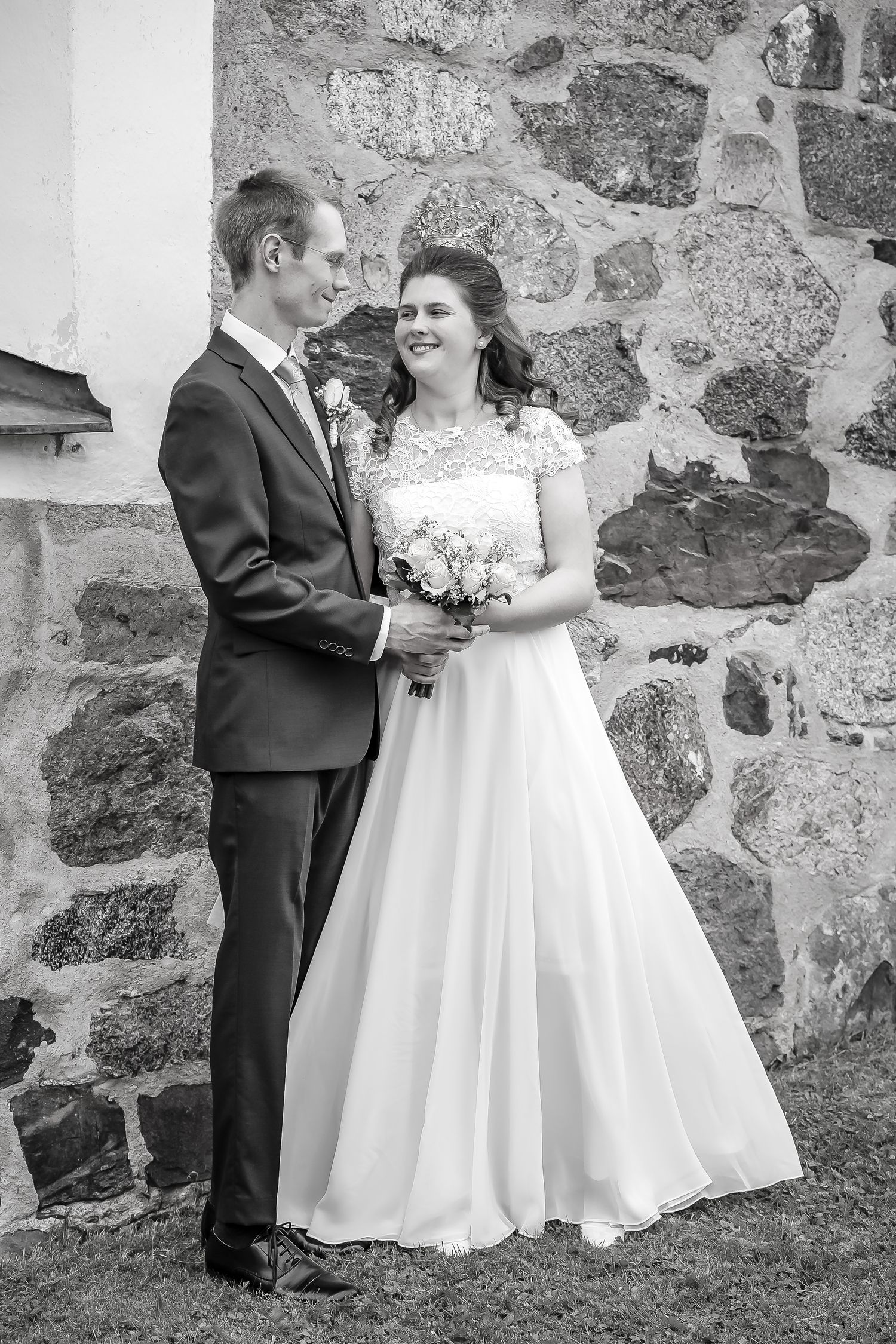 Black and white photo of a smiling bride holding bouquet and groom in suit standing by stone wall.