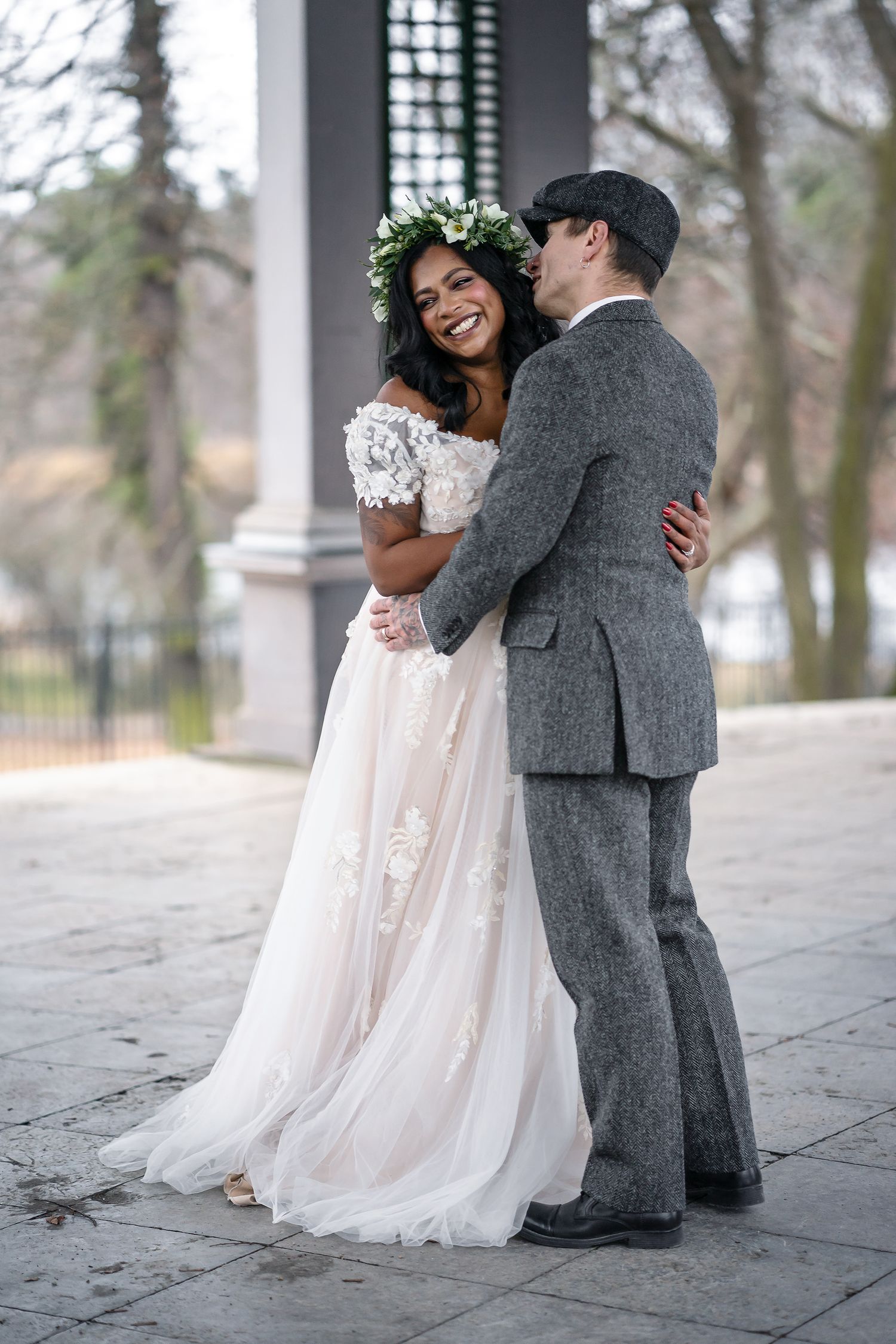 Smiling bride in floral crown and lace gown embraces groom in gray tweed suit outdoors.
