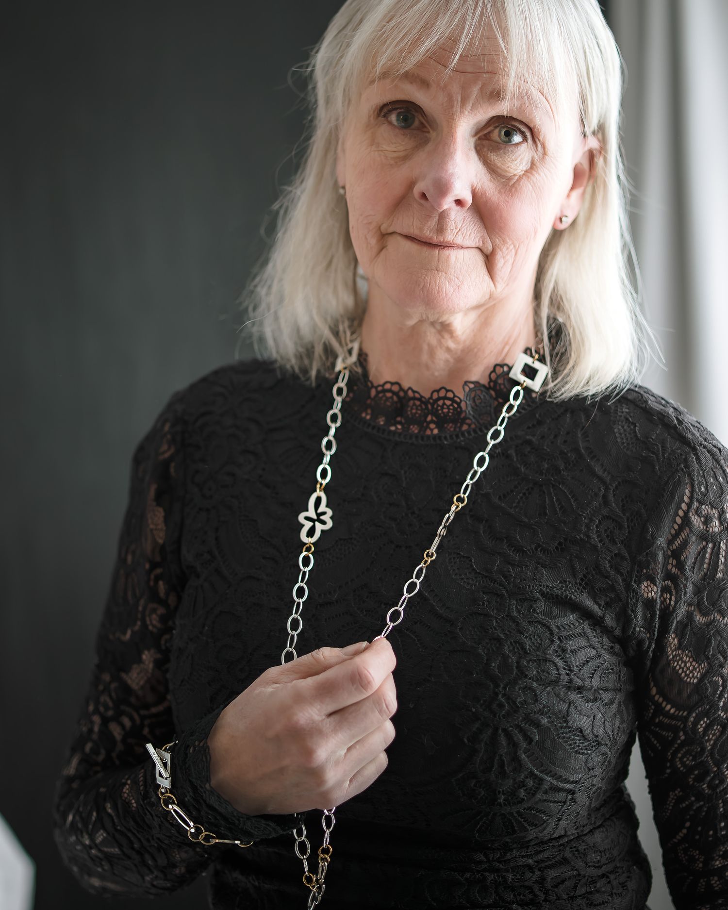 Someone wearing a black lace top and silver chain necklace poses against a muted background.
