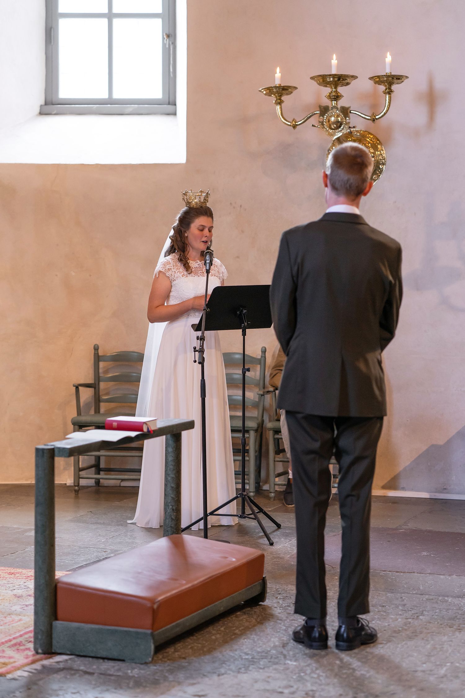 Bride in white dress reads vows at microphone stand while groom in gray suit listens during indoor wedding ceremony.