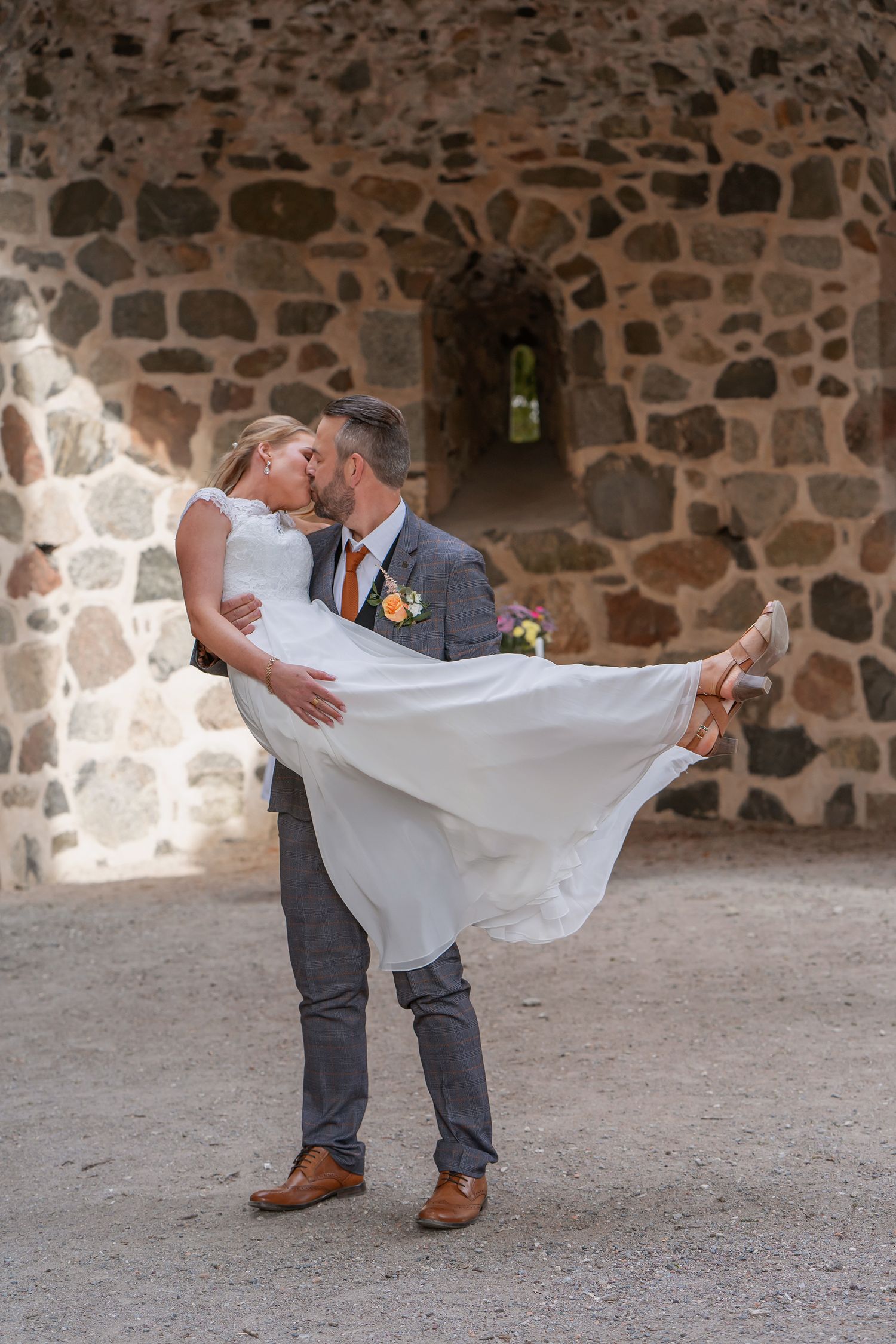 Groom lifting bride in white dress, sharing a kiss in front of a rustic stone wall archway.