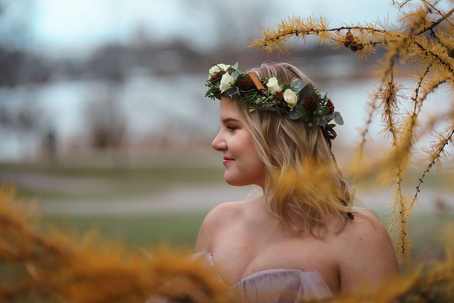 Young woman wearing a floral crown with white flowers poses outdoors among golden autumn foliage near a lake.