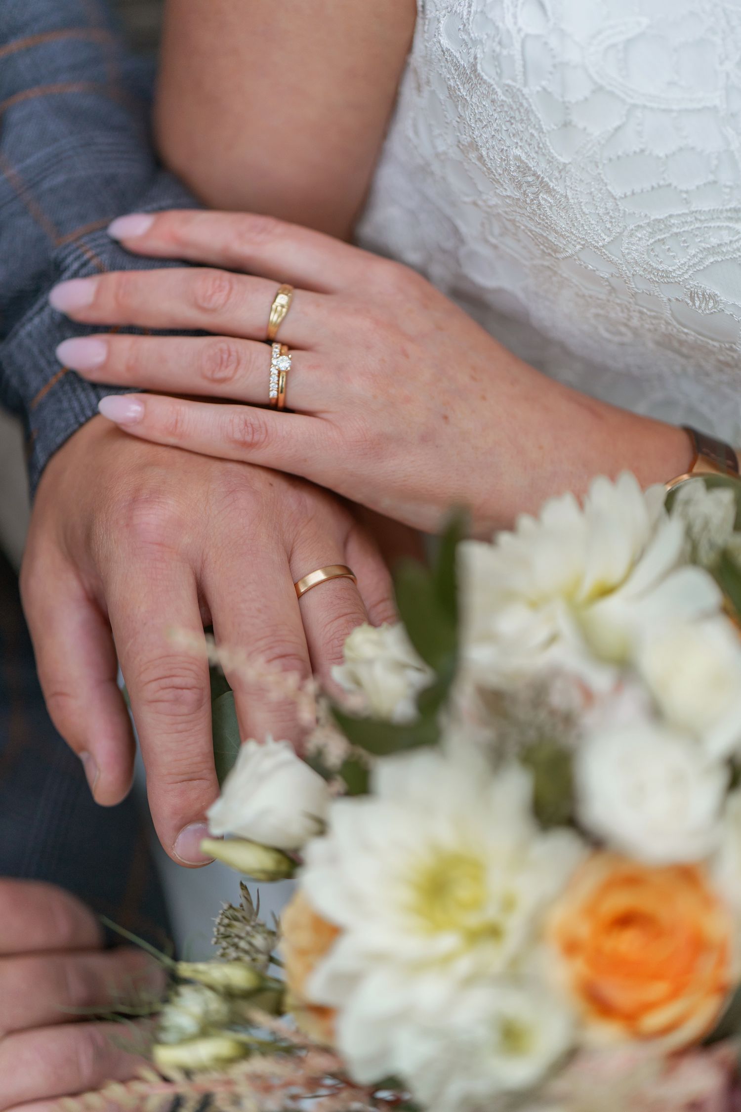 Bride and groom hands with gold wedding rings resting on a bridal bouquet of white and orange flowers.