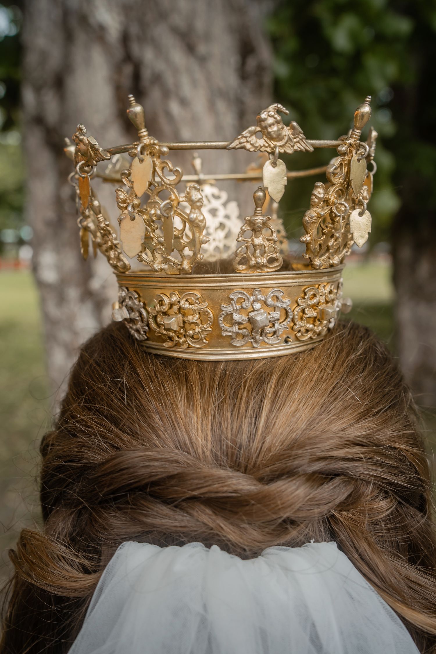 Back view of a woman wearing an ornate gold crown with heart charms in her braided updo, outdoors.