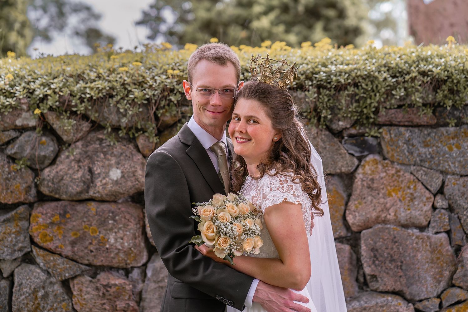 A smiling bride holding a bouquet of roses poses with her groom against a rustic stone wall covered in yellow lichen.