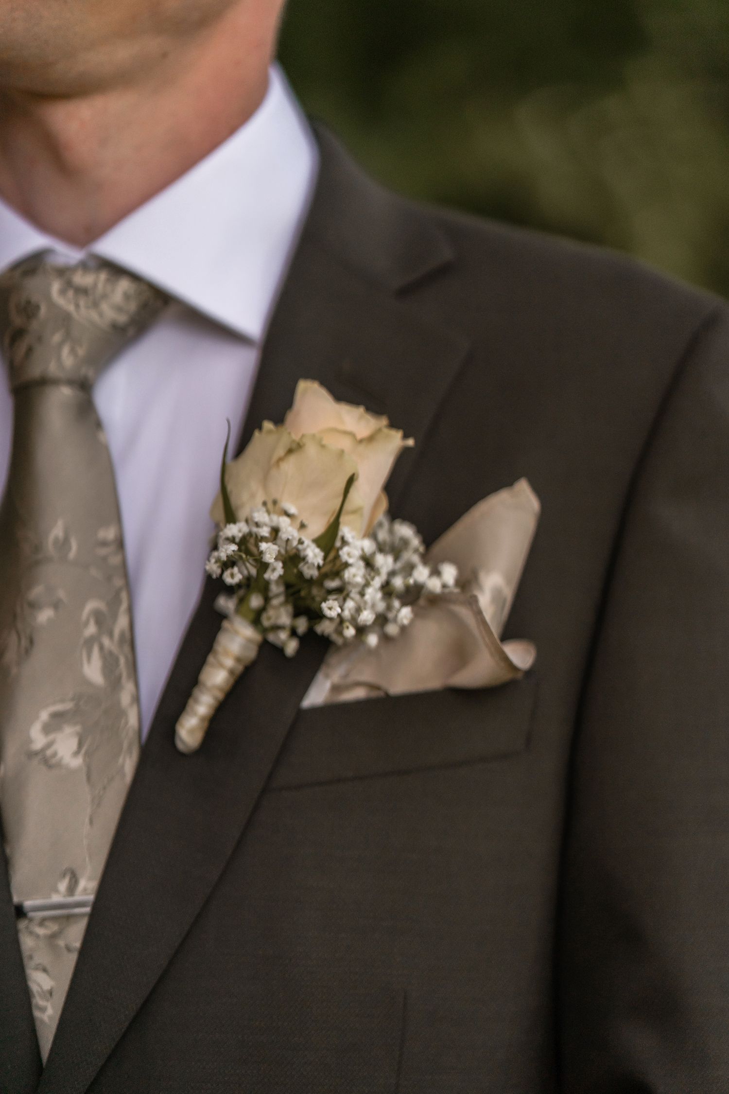 Man in gray suit with floral tie, cream rose boutonniere and matching pocket square at a formal event.