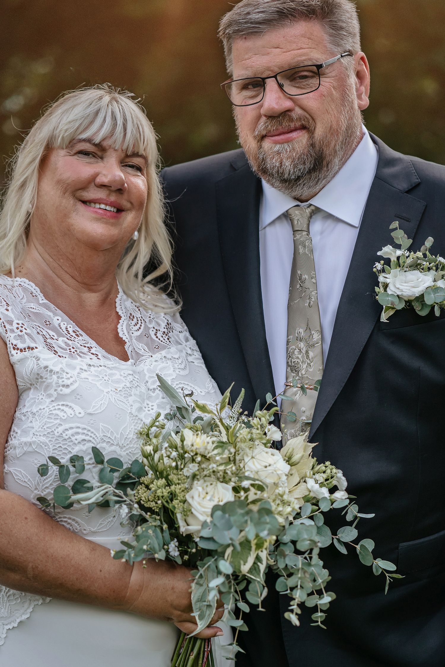Smiling bride in white lace dress holding bouquet with groom in dark suit and floral tie at outdoor wedding ceremony.
