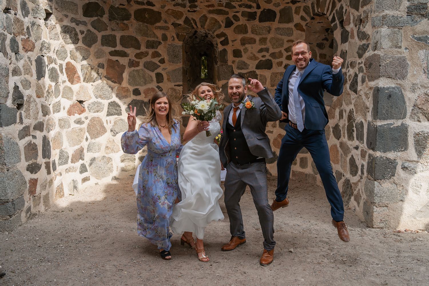 Joyful wedding party celebrates with jumping and cheering in a rustic stone archway venue.
