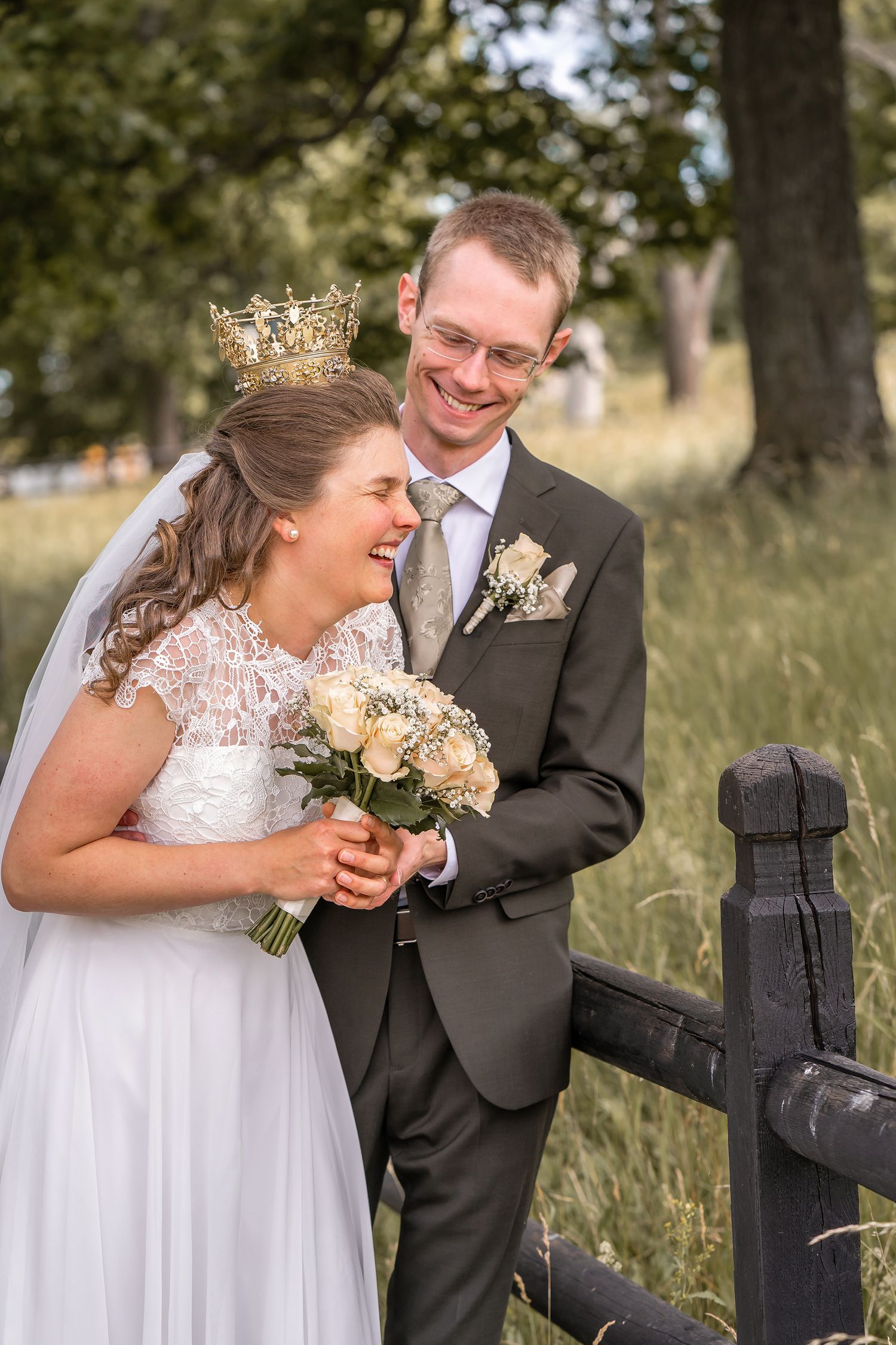 Bride wearing crown and veil holds bouquet while laughing with groom in gray suit near wooden fence outdoors.