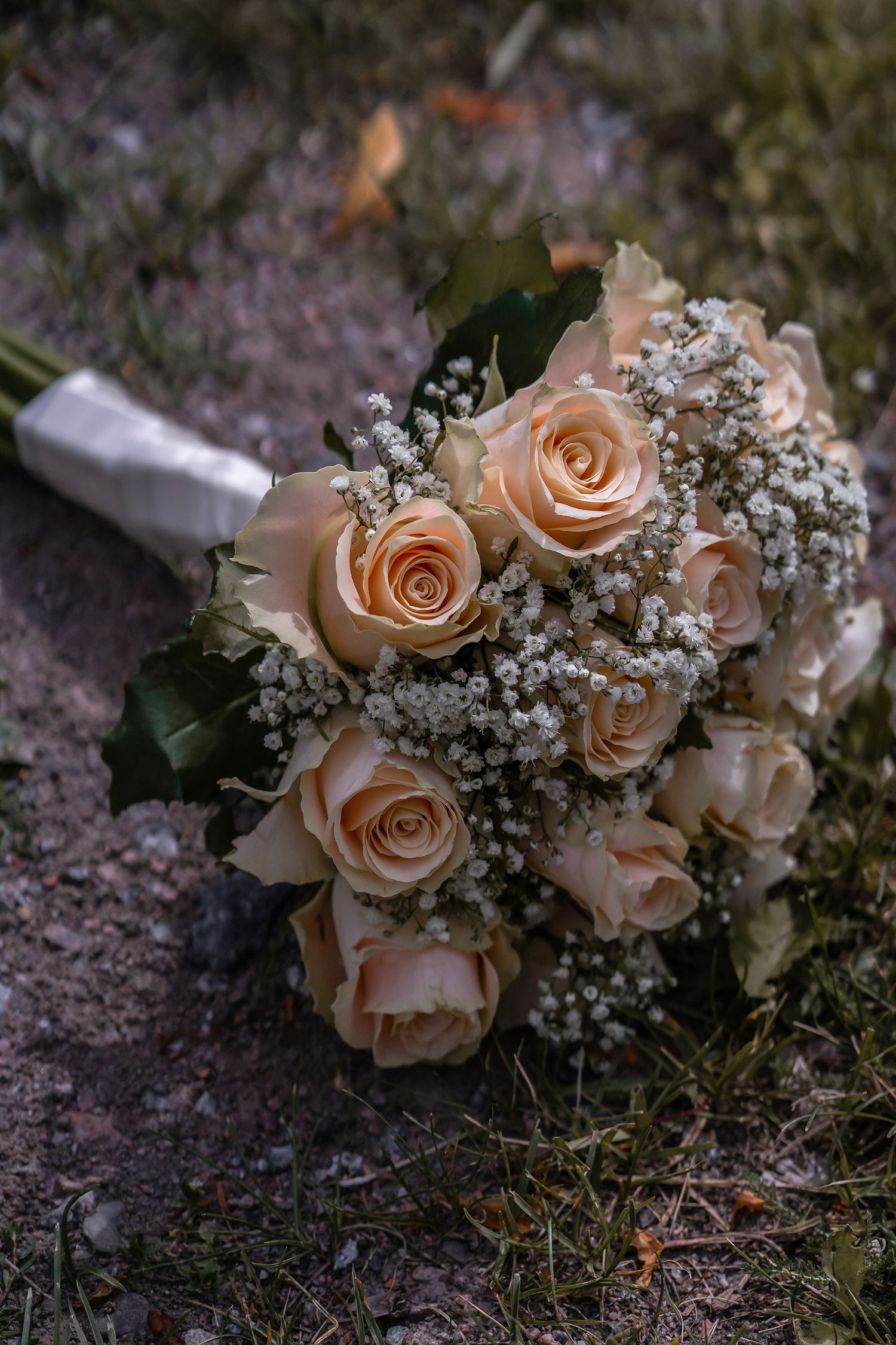Elegant bridal bouquet of peach roses and white baby's breath with ribbon, resting on natural ground.
