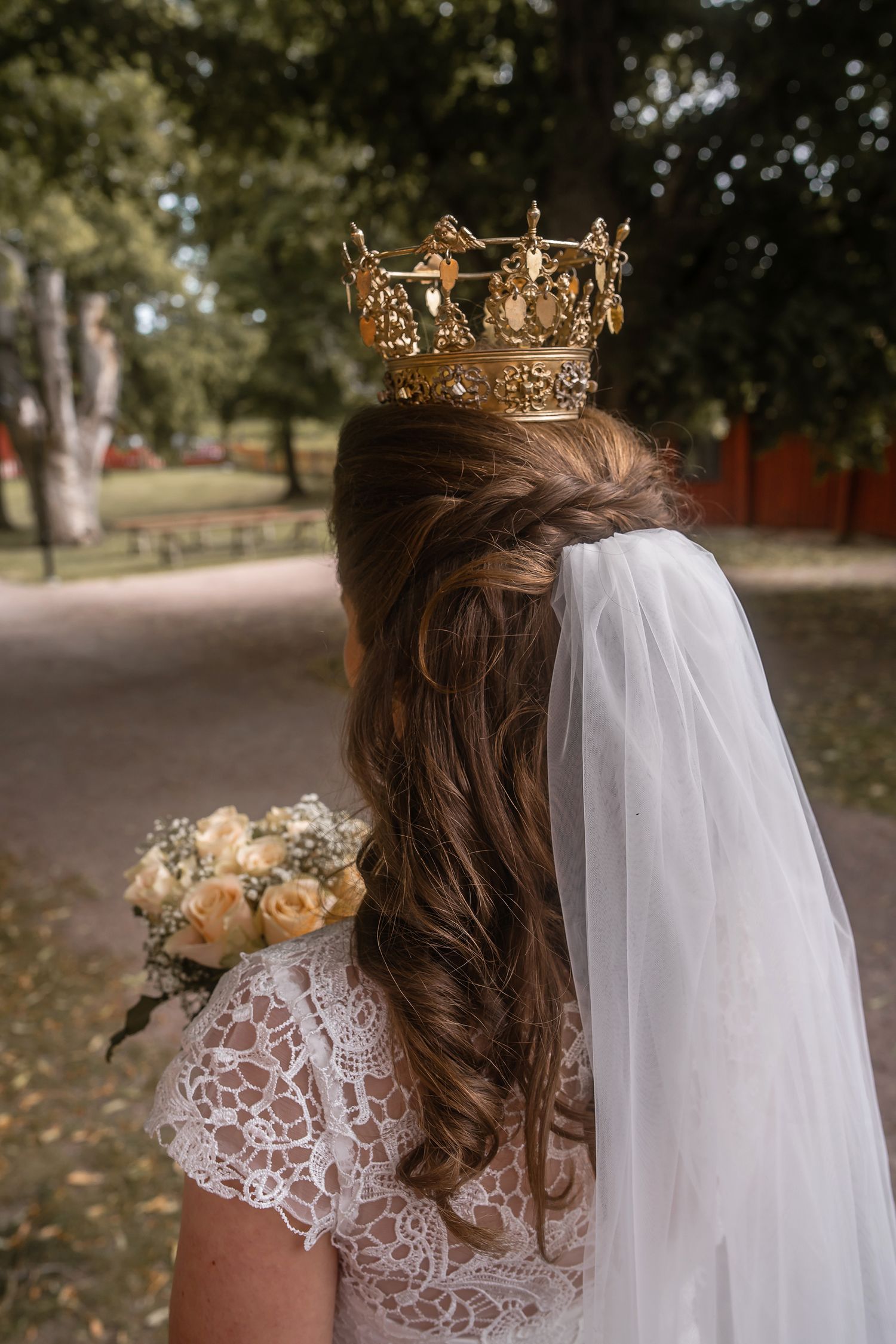 Bride with golden crown, white veil, and lace dress holding bouquet of roses in a park setting.