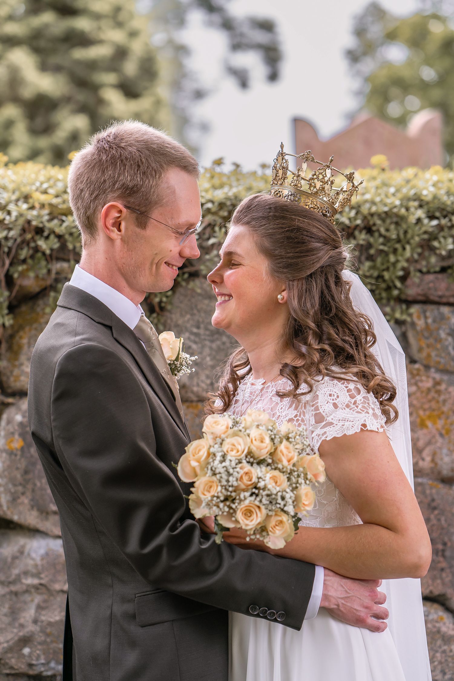 A smiling bride wearing a crown holds a bouquet of roses while embracing a groom in a gray suit outdoors.