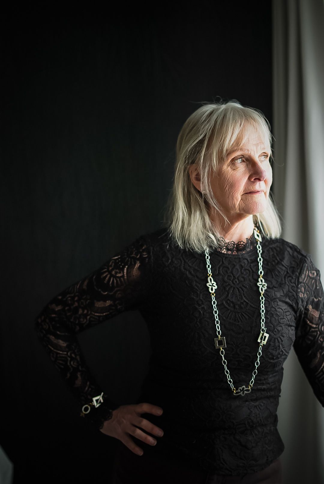A person wearing a black lace dress and silver jewelry poses against a window in dramatic lighting.