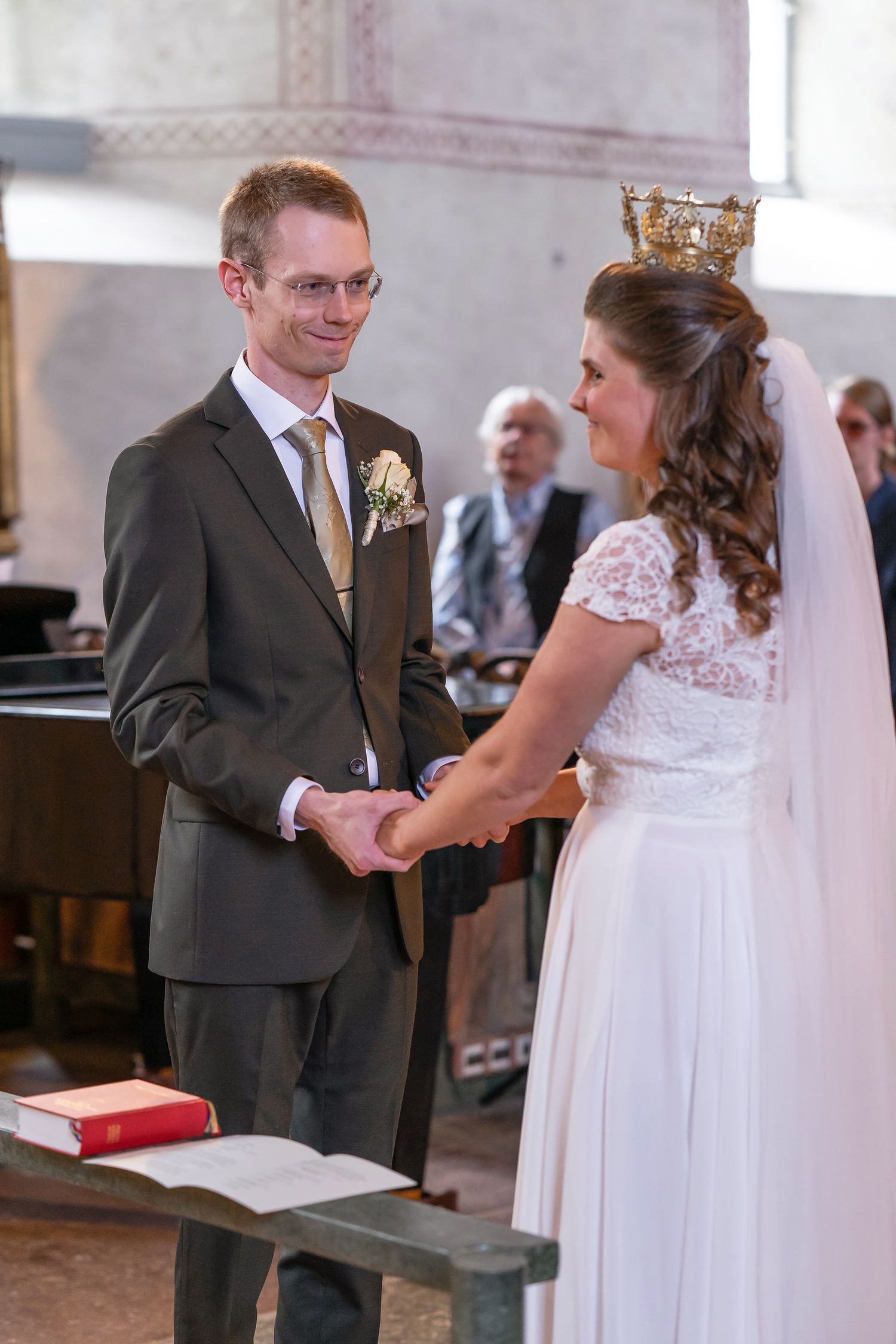 Bride and groom holding hands at church altar during wedding ceremony, bride wearing crown and white lace dress.