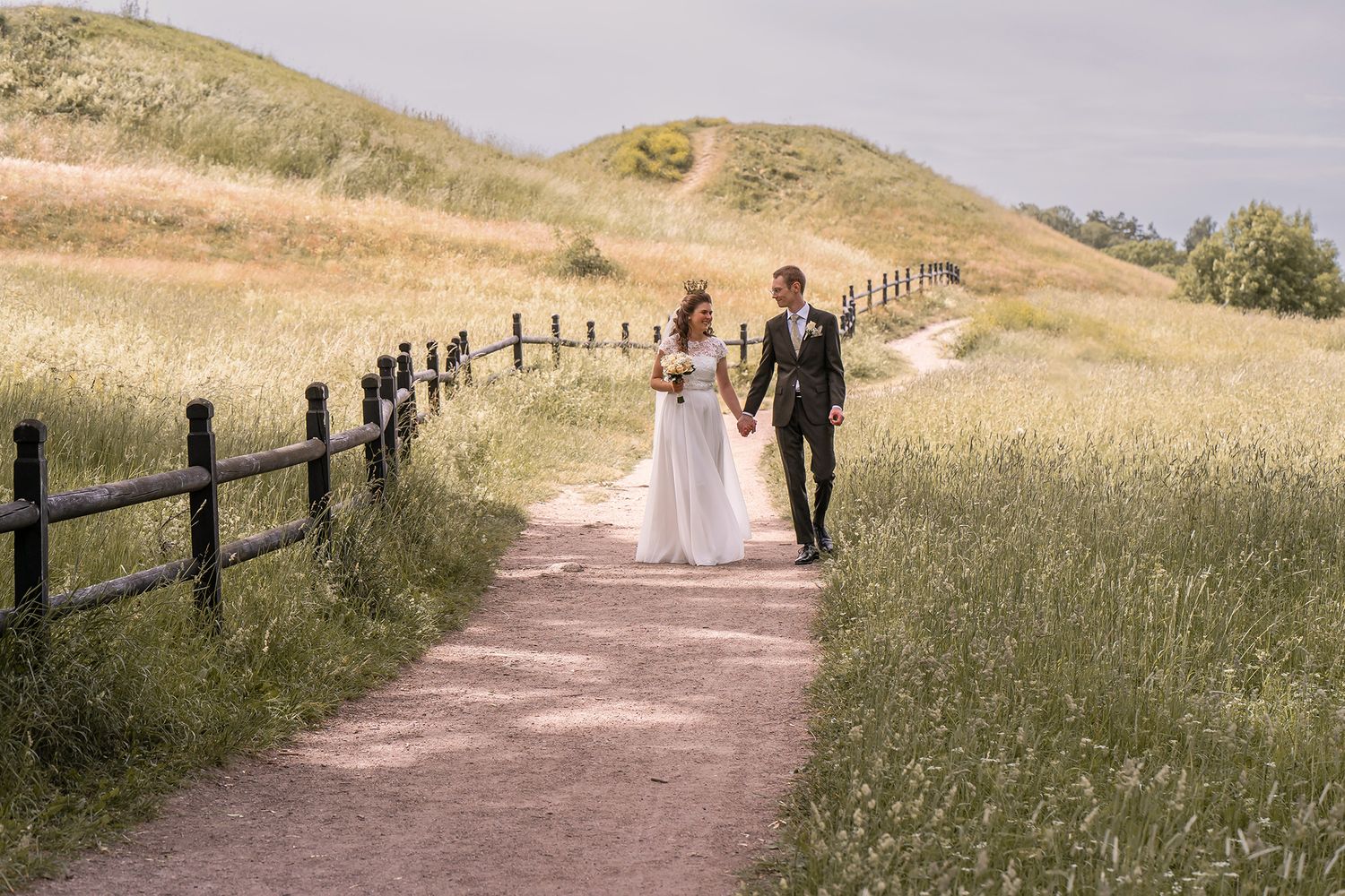 A bride and groom walk hand in hand along a scenic dirt path surrounded by lush green fields and a wooden fence.
