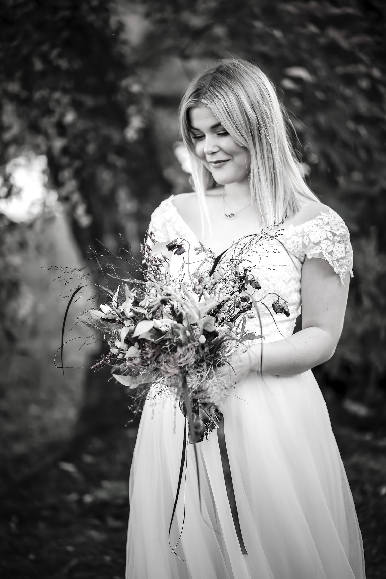 Black and white photo of a blonde bride in a white lace gown holding a wildflower bouquet, smiling softly outdoors.