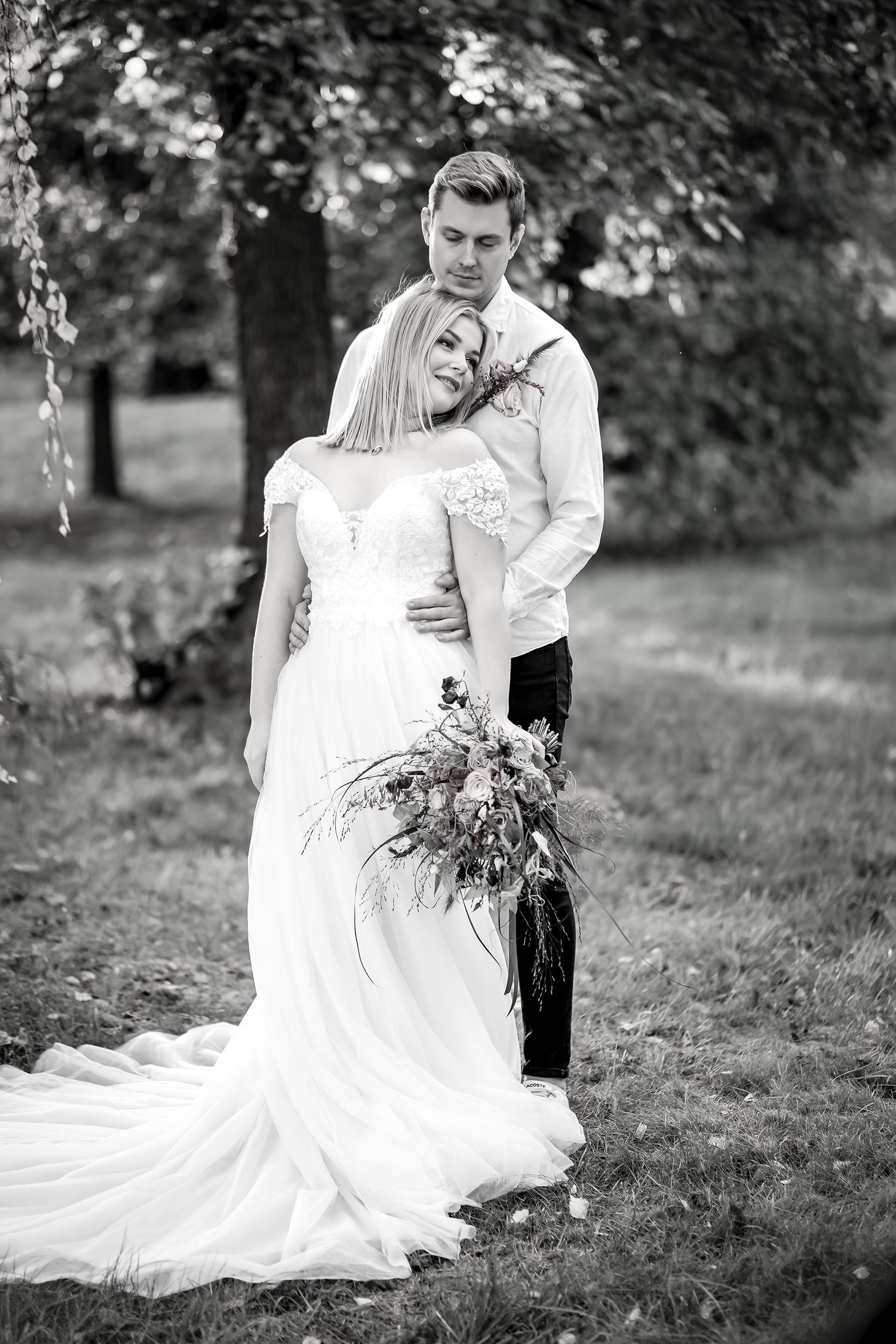 Black and white photo of a bride and groom embracing outdoors, bride holding a bouquet, groom in white shirt.