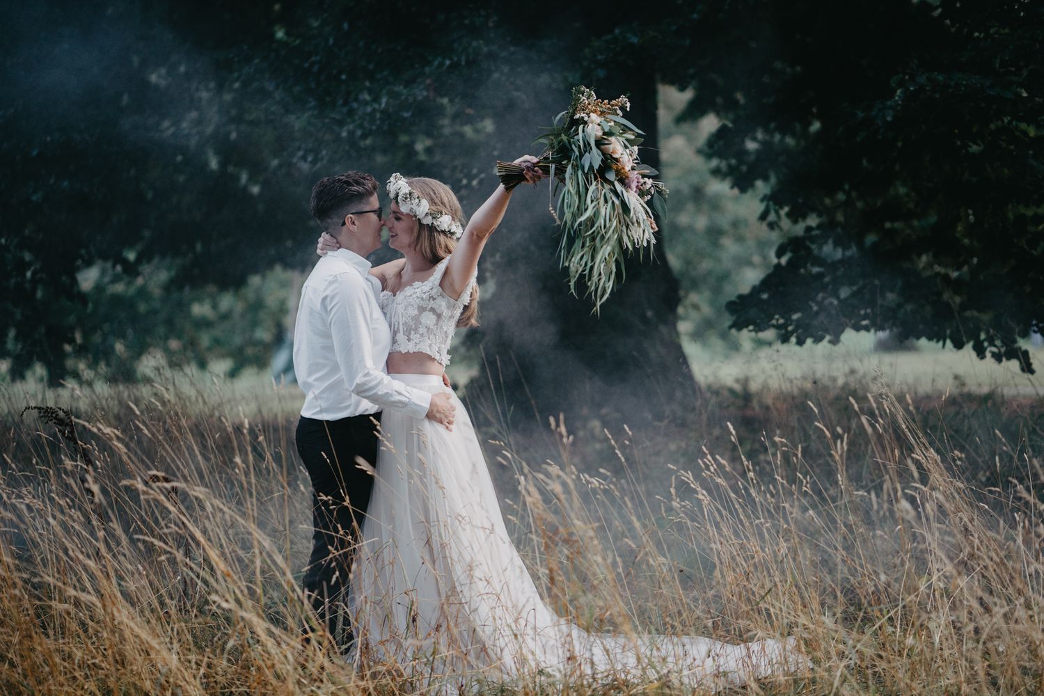 Bride and groom share joyful moment in golden field, bride holding lush bouquet aloft on wedding day.