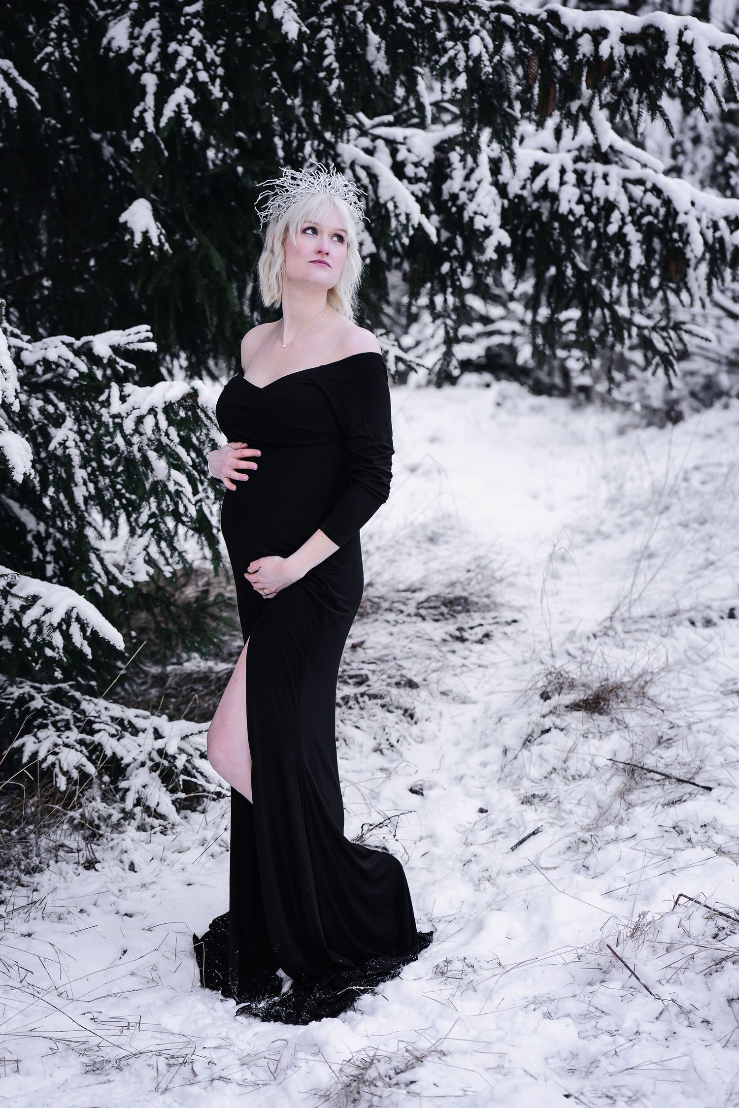 Gravid kvinna i en elegant svart off-shoulder-klänning med krona, poserar i en snötäckt vinterskog.
